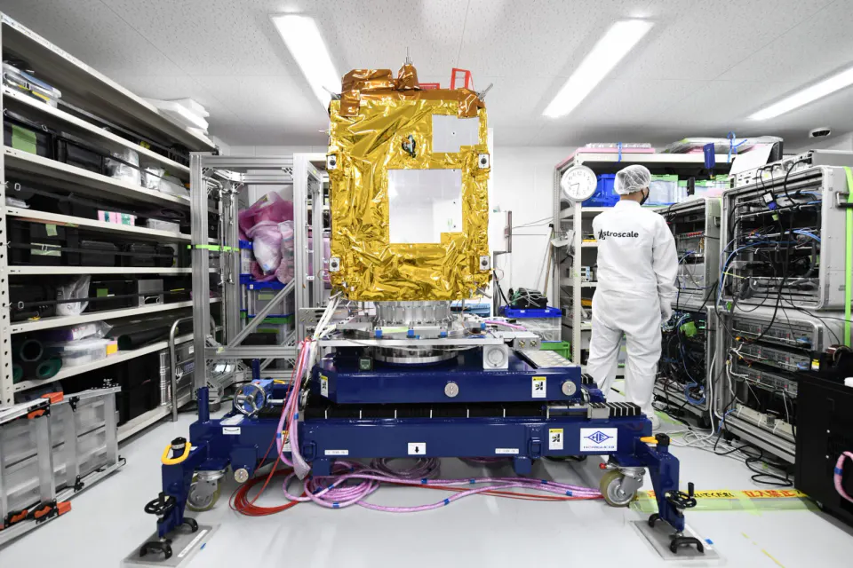 白い作業着の技術者が立つクリーンルームで、金色の断熱材に包まれた箱型の衛星が台の上に載っている