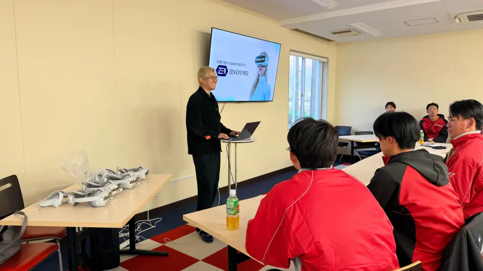 Fuma Hayashi stands at a laptop in front of a monitor, explaining ZEN EYE Pro to Honda baseball players in red team jackets