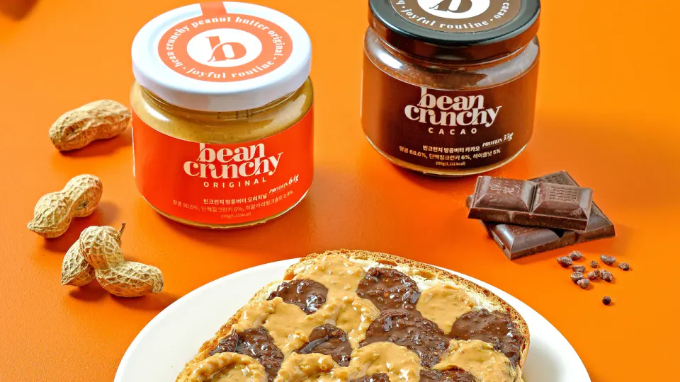 オレンジ色の背景に並ぶ、BeanCrunchyのオリジナルとカカオのピーナッツバターの瓶。手前にピーナッツバターとチョコレートを塗ったパン